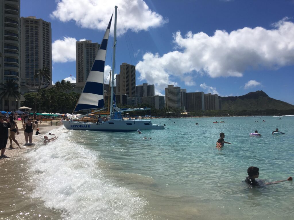 ワイキキビーチの浅瀬で泳ぐ人たちと、砂浜に上げられたストライプ柄のカタマラン。奥にダイヤモンドヘッドとホテル群