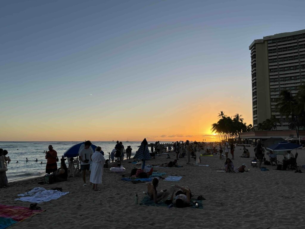 夕日が沈むワイキキビーチ。砂浜に人が集まり、ヤシの木とホテルの向こうに日が沈んでいく